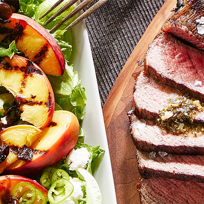 Two plates of food: one a plate of mixed greens topped with  grilled peaches, the other a sliced grilled steak topped with chimichurri sauce. 