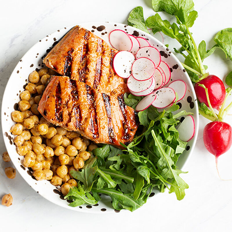 A bowl filled with salad consisting of garbanzo beans, arugala, and thinly sliced radishes and topped with two filets of grilled salmon.